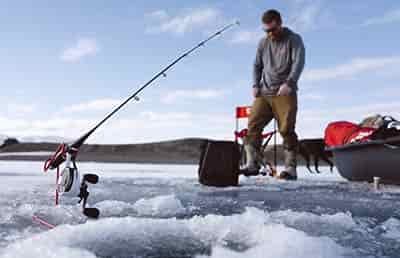 man ice fishing with pole staked in ice hands in pocket waiting on fish bite