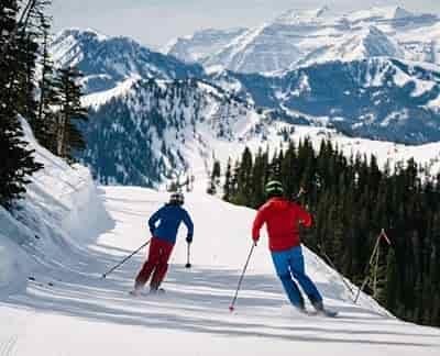 couple skiing down groomed run snow covered mountain in background
